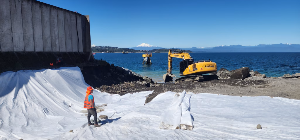 Excavadora LiuGong trabajando en la costa durante la ampliación del Puerto Punta Caullahuapi, con geotextil blanco en primer plano y el mar y montañas al fondo.